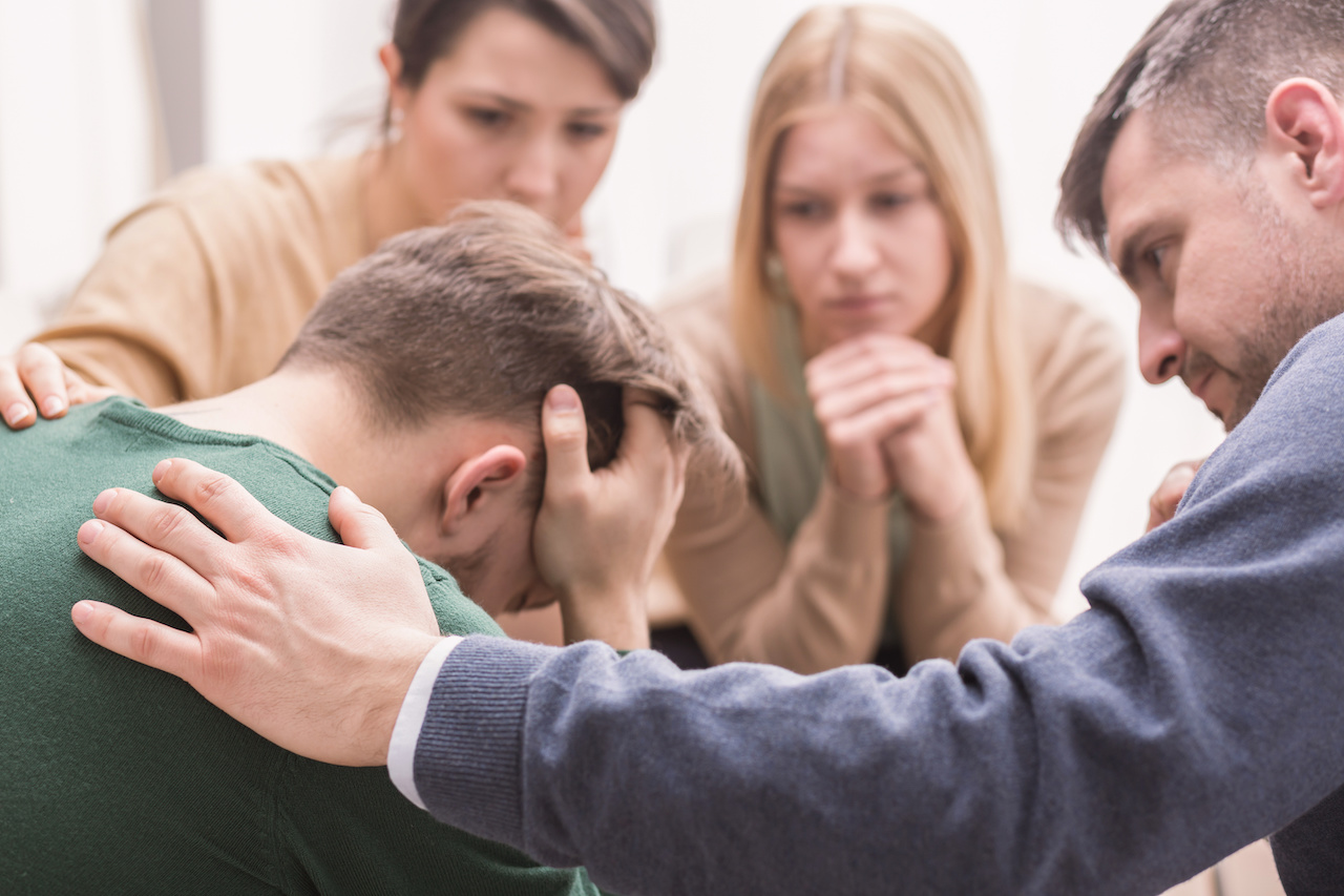 Close-up of a devastated young man holding his head in his hands and family supporting him during group therapy jpeg