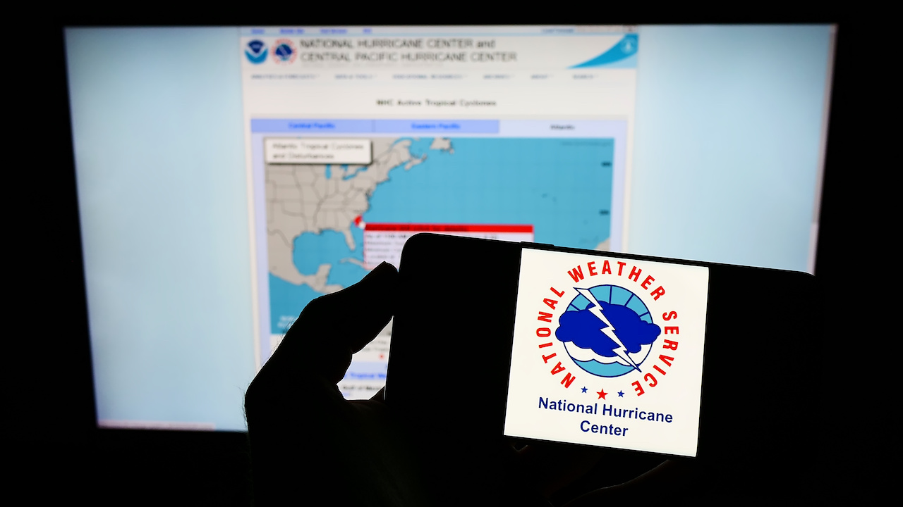 Person holding smartphone with logo of National Hurricane Center (NHC) on screen in front of website jpeg